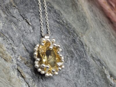 Oval, golden yellow gemstone pendant with a silver bezel and beaded setting, resting on a grey stone surface.