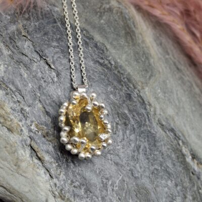 Oval, golden yellow gemstone pendant with a silver bezel and beaded setting, resting on a grey stone surface.