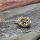 Oval, golden yellow gemstone pendant with a silver bezel and beaded setting, resting on a grey stone surface.