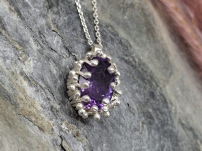 Oval, dark purple gemstone pendant with a silver bezel and beaded setting, resting on a grey stone surface.