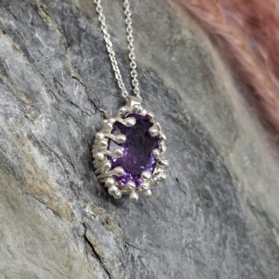 Oval, dark purple gemstone pendant with a silver bezel and beaded setting, resting on a grey stone surface.