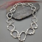 Textured silver necklace with interlocking, organic rings and a toggle clasp, resting on a grey stone surface.