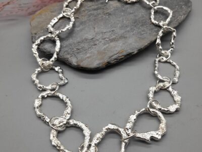 Textured silver necklace with interlocking, organic rings and a toggle clasp, resting on a grey stone surface.