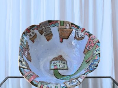 This food-safe bowl features playful, colourful houses with a winding path. The light blue backdrop and raised details add texture. It was handcrafted and signed by Dorienne and is perfect for serving or decoration.